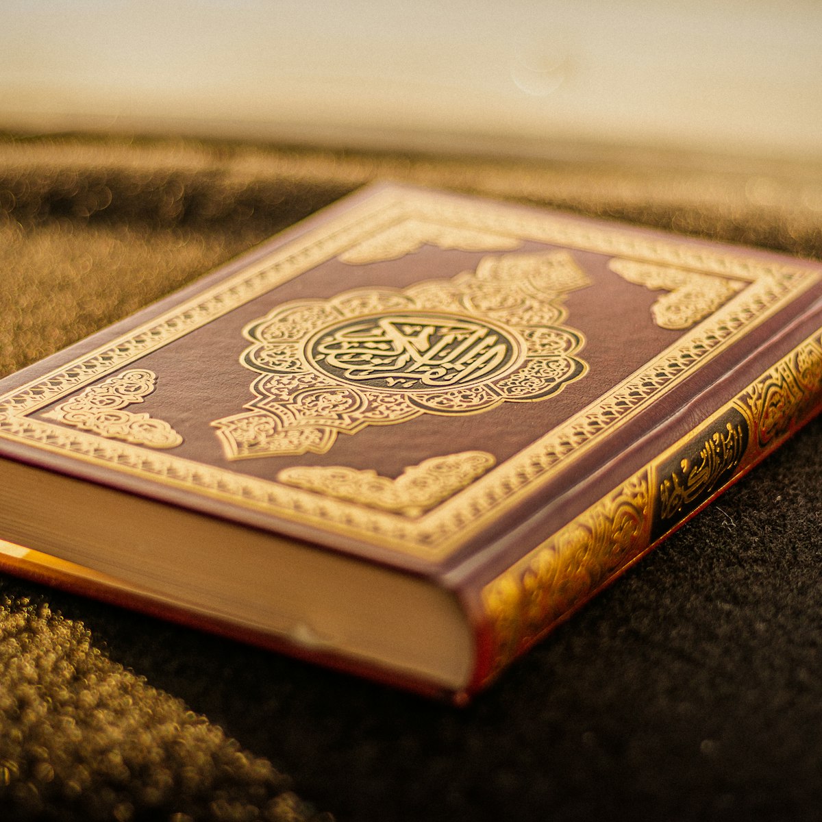 A gold-embossed leather-bound Qur'an resting on sand at golden hour
